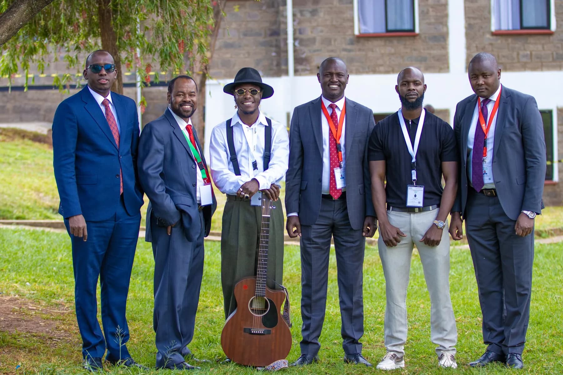 Six men in suits and conference lanyards standing together on a lawn, one holding an acoustic guitar