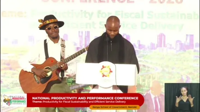 Two performers at the conference podium, one playing an acoustic guitar, shown in a television broadcast