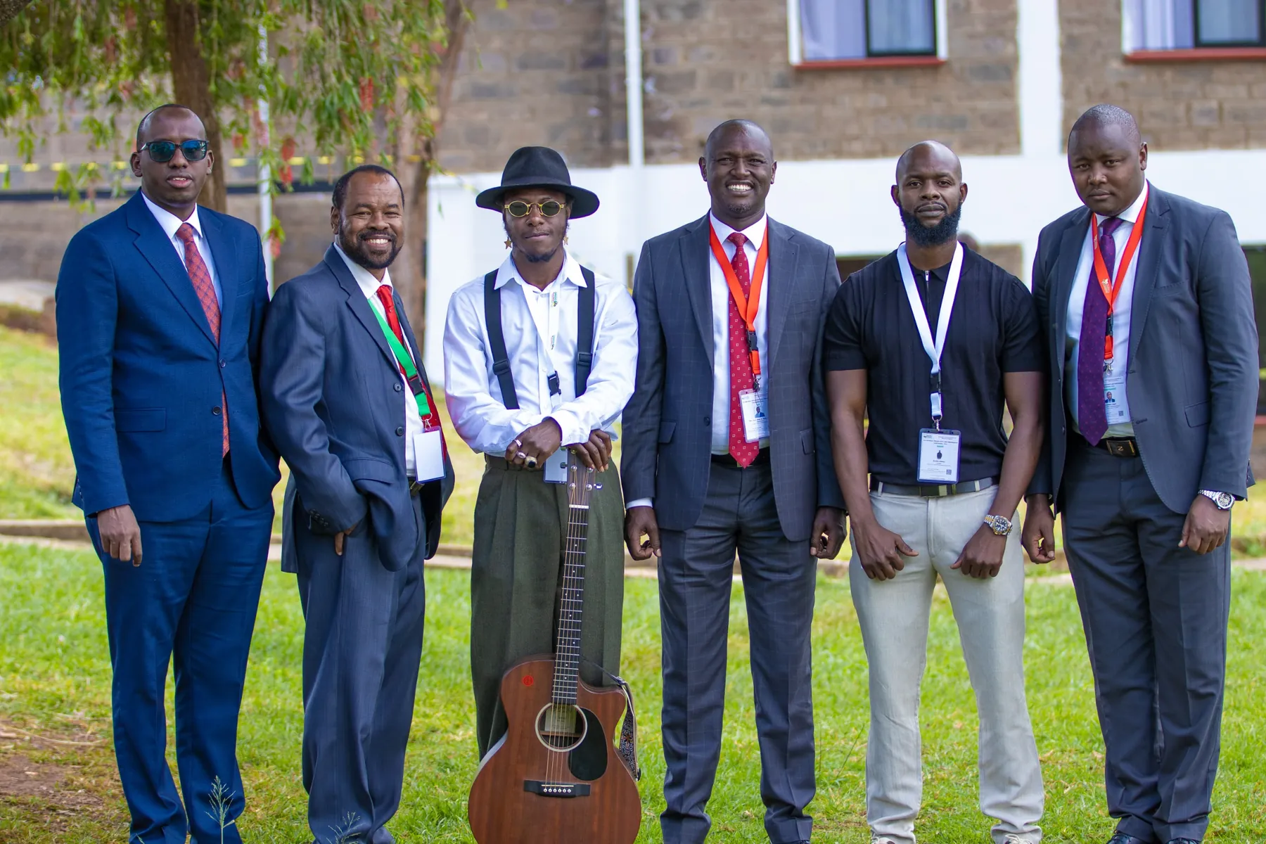 Six men photographed together on a lawn outside a conference venue, five in suits with delegate lanyards and one holding an acoustic guitar