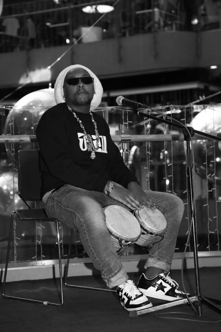 A drummer seated at a microphone with a pair of hand drums held between his knees