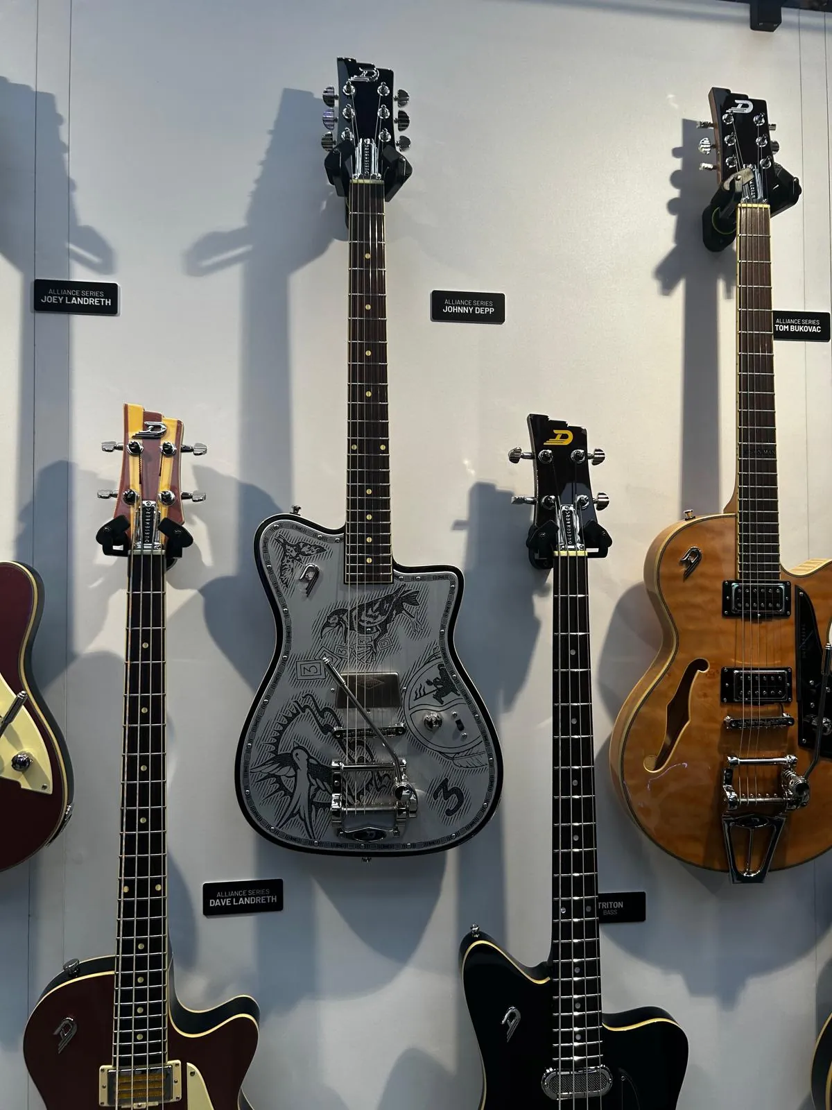 A wall of electric guitars and basses on display, each with a manufacturer's name plate beneath it