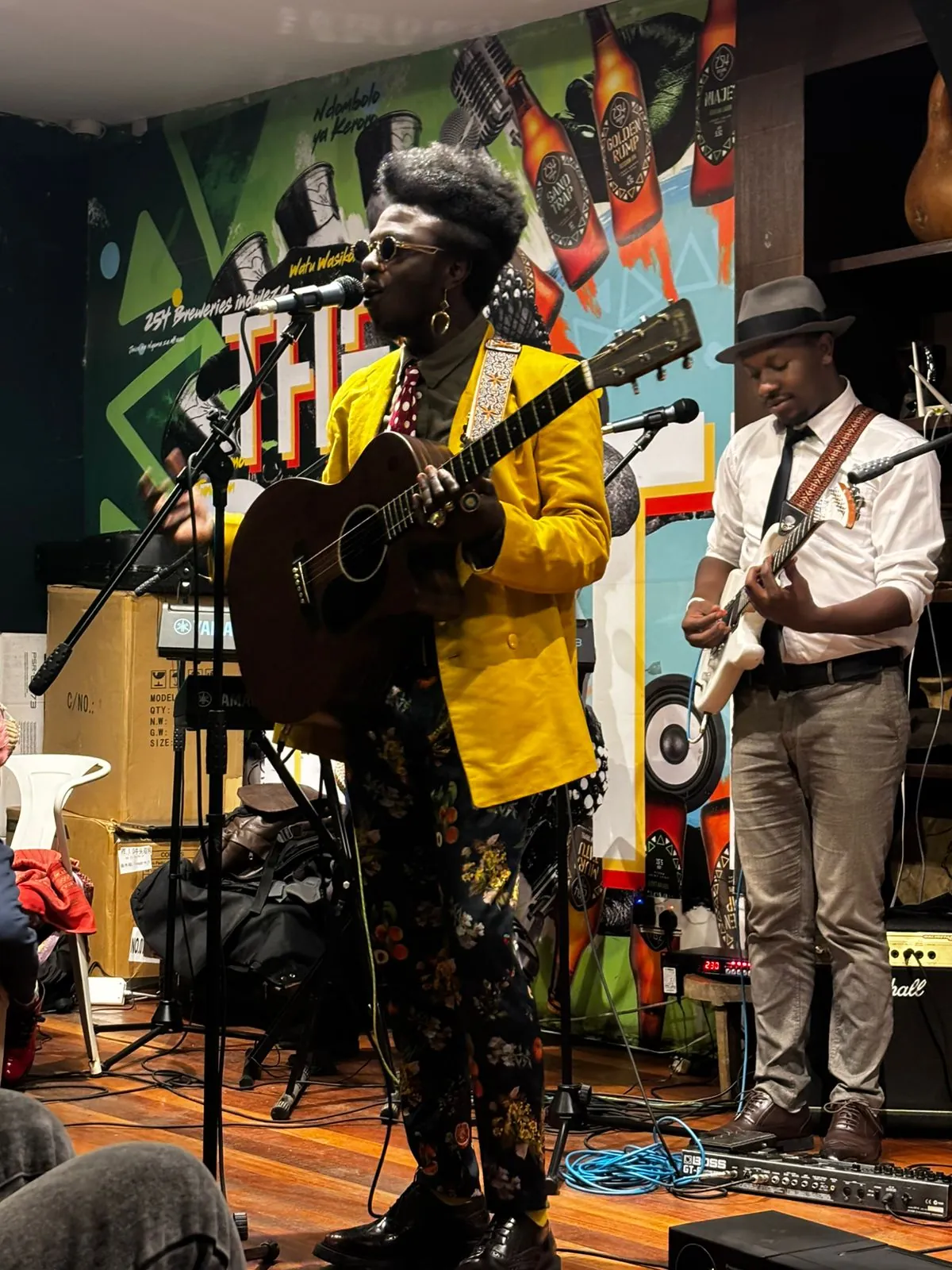 Two musicians performing together on a small stage, one on acoustic guitar at the microphone and one on electric guitar