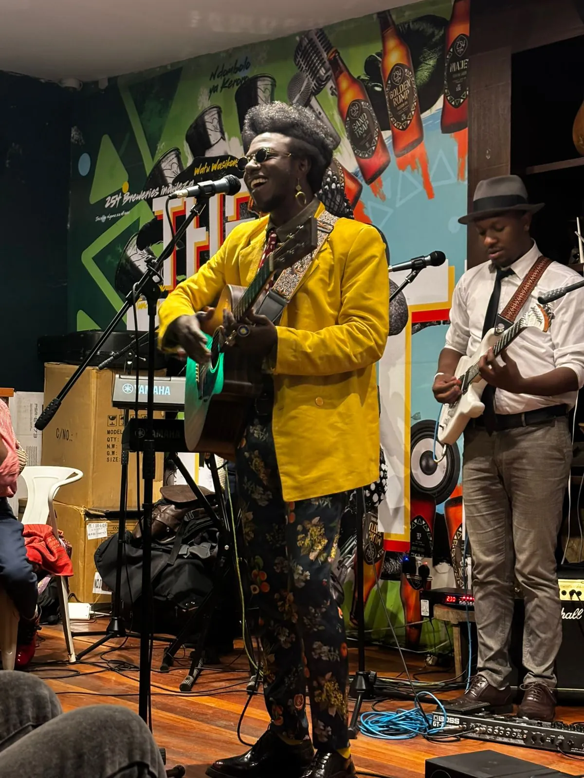 A singer in a yellow jacket playing an acoustic guitar at a microphone, a guitarist beside him