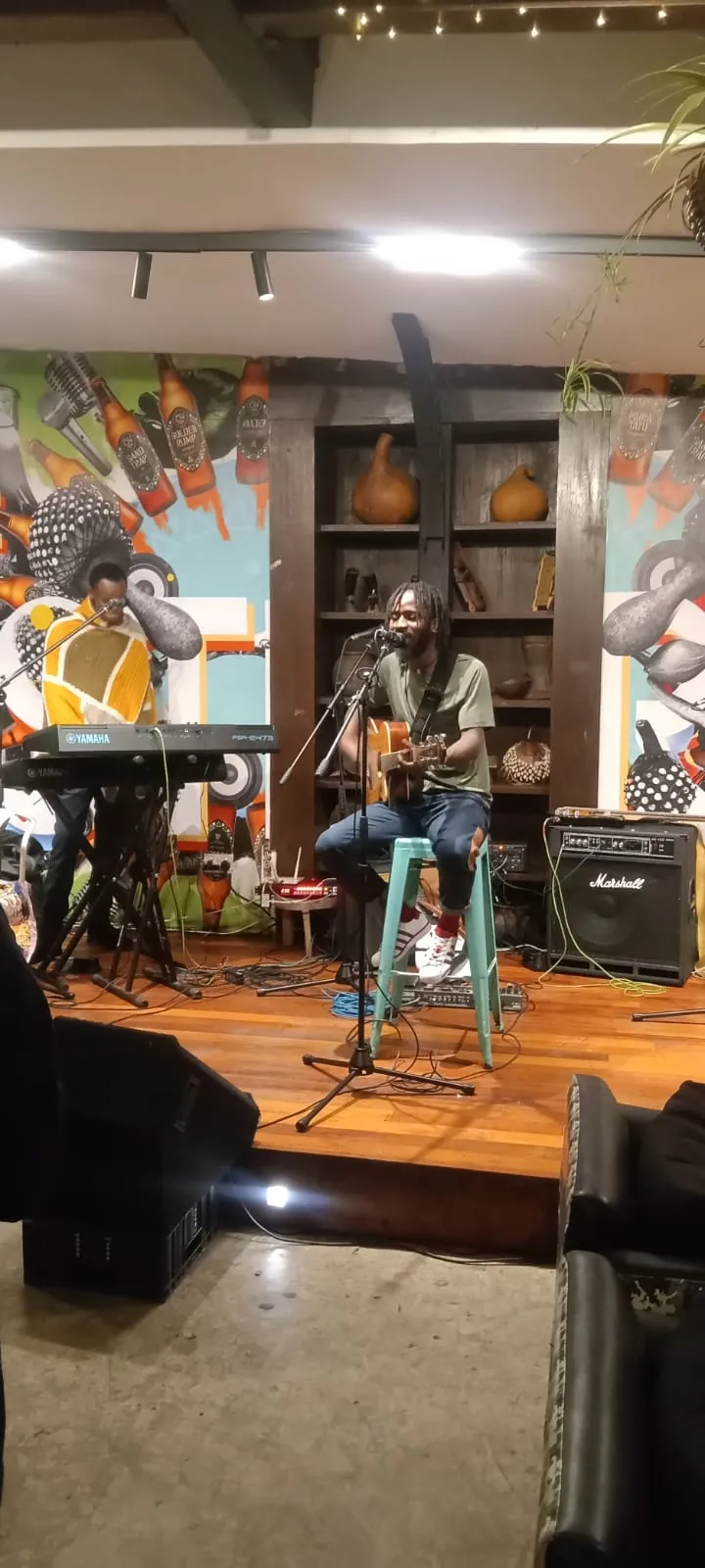 A solo performer seated on a stool with an acoustic guitar, a keyboard player behind him