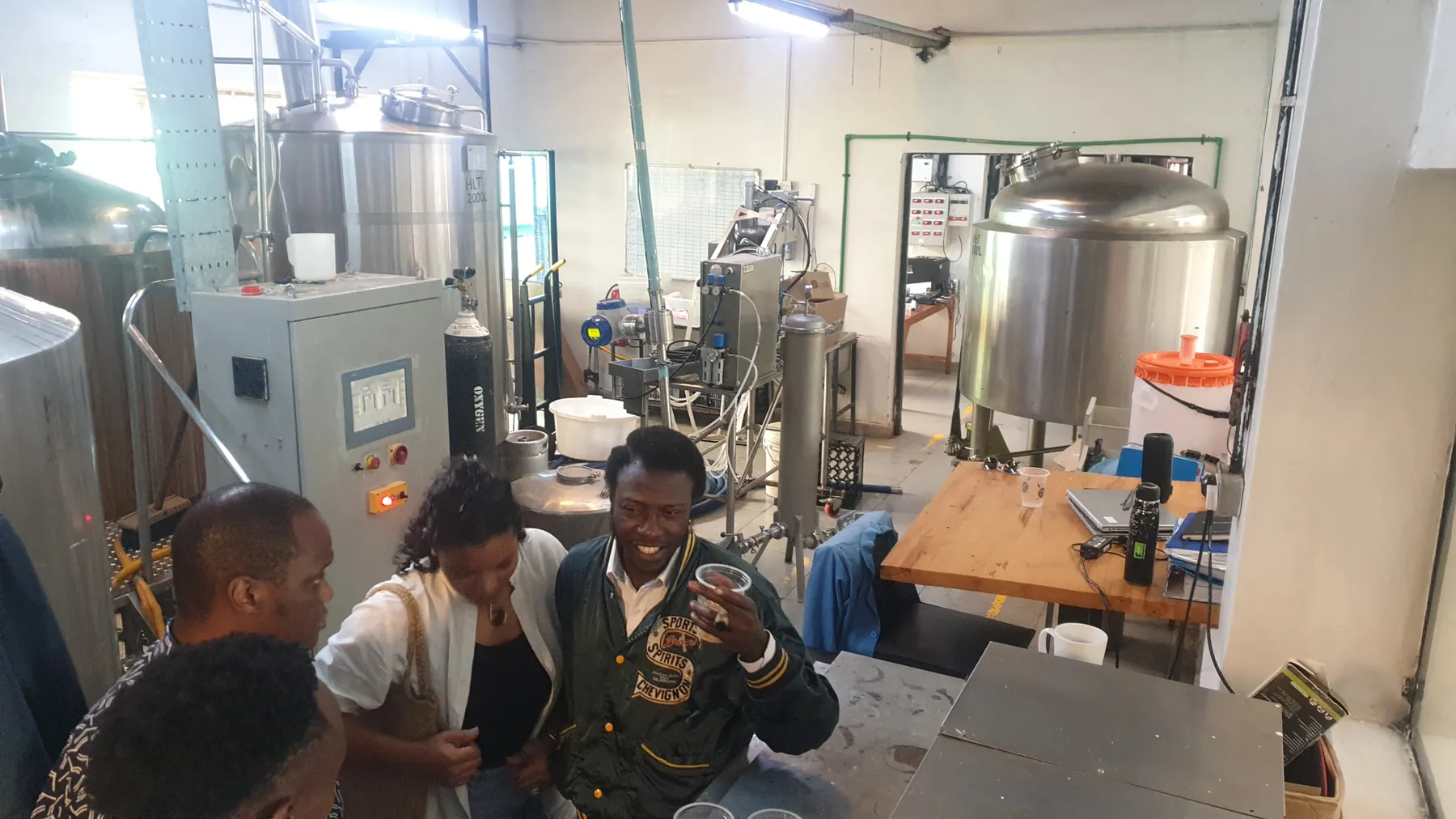 A wider view of the same brewery floor, tanks and control panels behind a group in conversation